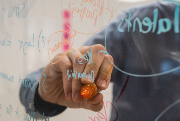Una mano sostiene un rotulador naranja tras una pizarra de cristal, donde hay anotaciones hechas en color azul y naranja.