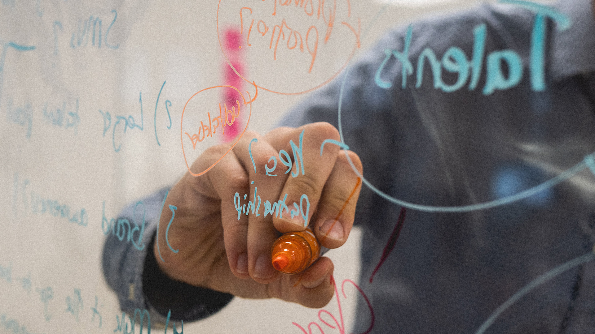 Una mano sostiene un rotulador naranja tras una pizarra de cristal, donde hay anotaciones hechas en color azul y naranja.